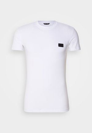 Cămașă albă din bumbac, cu decolteu rotund și mâneci scurte. Include un mic accent cu etichetă neagră în zona stângă a pieptului.