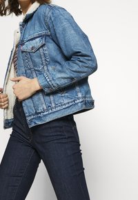 Jeansjacka i ljusblått med en mjuk, krämfärgad foder. Har två bröstfickor och knappstängningar, uppvisad tillsammans med mörka jeans.