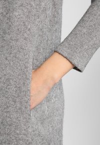 Ręka wsunięta w boczną kieszeń szarego wełnianego swetra z długimi rękawami.