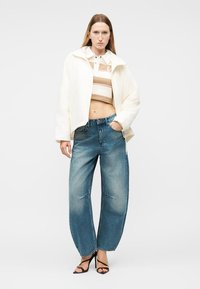 Μία γυναίκα στέκεται φορώντας λευκό κοντό σακάκι, μπεζ ριγέ κοντό τοπ, τζιν παντελόνι με φαρδιά μπατζάκια και μαύρες ψηλοτάκουνες σανδάλια, μπροστά σε απλό φόντο.