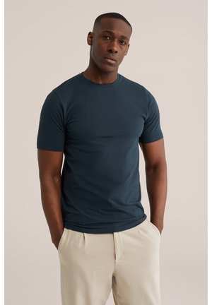 Muž stojí s rukami v vreckách, oblečený v tmavomodrom tričku s krátkym rukávom a svetlo béžových nohaviciach na jednofarebnom pozadí.