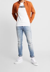 Camisa de botones naranja llevada sobre una camiseta gráfica blanca, combinada con unos vaqueros de mezclilla claros y desgastados y zapatillas blancas.