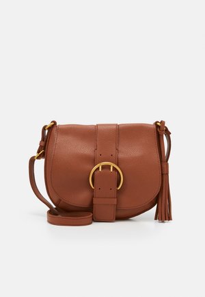 Sac bandoulière en cuir marron avec une grande boucle circulaire dorée, une sangle réglable et un détail pompon sur un fond clair.