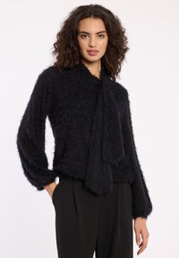 Schwarzer, flauschiger Strickpullover mit Schal-Detail und langen Ärmeln, in lockerer Passform und strukturierter Verarbeitung, kombiniert mit schwarzen Hosen.