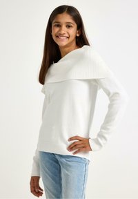 Maglione bianco con scollo a coste a spalle scoperte e maniche lunghe, abbinato a jeans azzurri. Tessuto morbido, vestibilità rilassata.