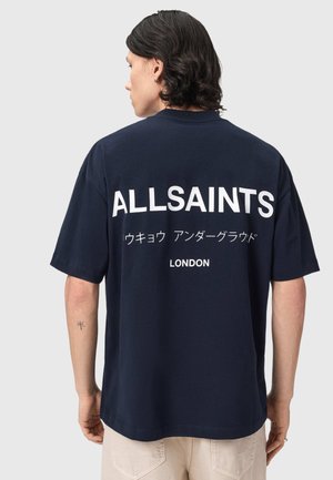 Person with shoulder-length dark hair wears a navy blue shirt with "ALLSAINTS" and Japanese characters printed on the back above "LONDON."