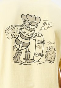 Geltona marškinėliai, kuriuose pavaizduotas animacinis bites cowboy skrybėlėje ir botose, laikantis riedlentę, šalia animacinio ežiuko. Juodos linijos dailės dizainas.