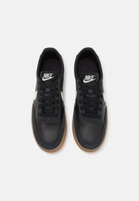 Baskets en cuir noir avec une semelle en caoutchouc beige, arborant un logo Nike blanc, des lacets noirs et une texture supérieure lisse.