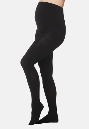 Pregnant person wearing black maternity tights standing sideways with one foot slightly raised against a plain light background.
