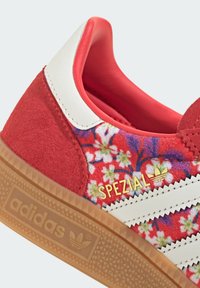 Zapatilla roja Adidas Spezial con patrón floral, rayas blancas, marca dorada y suela de goma en vista en primer plano que muestra detalles del talón y los laterales.