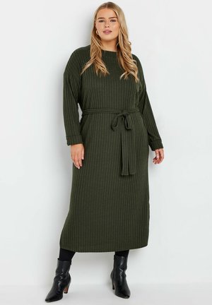 Robe longue verte à côtes avec des manches longues et une taille cintrée. Le tissu a une finition texturée. Portée avec des bottines noires.