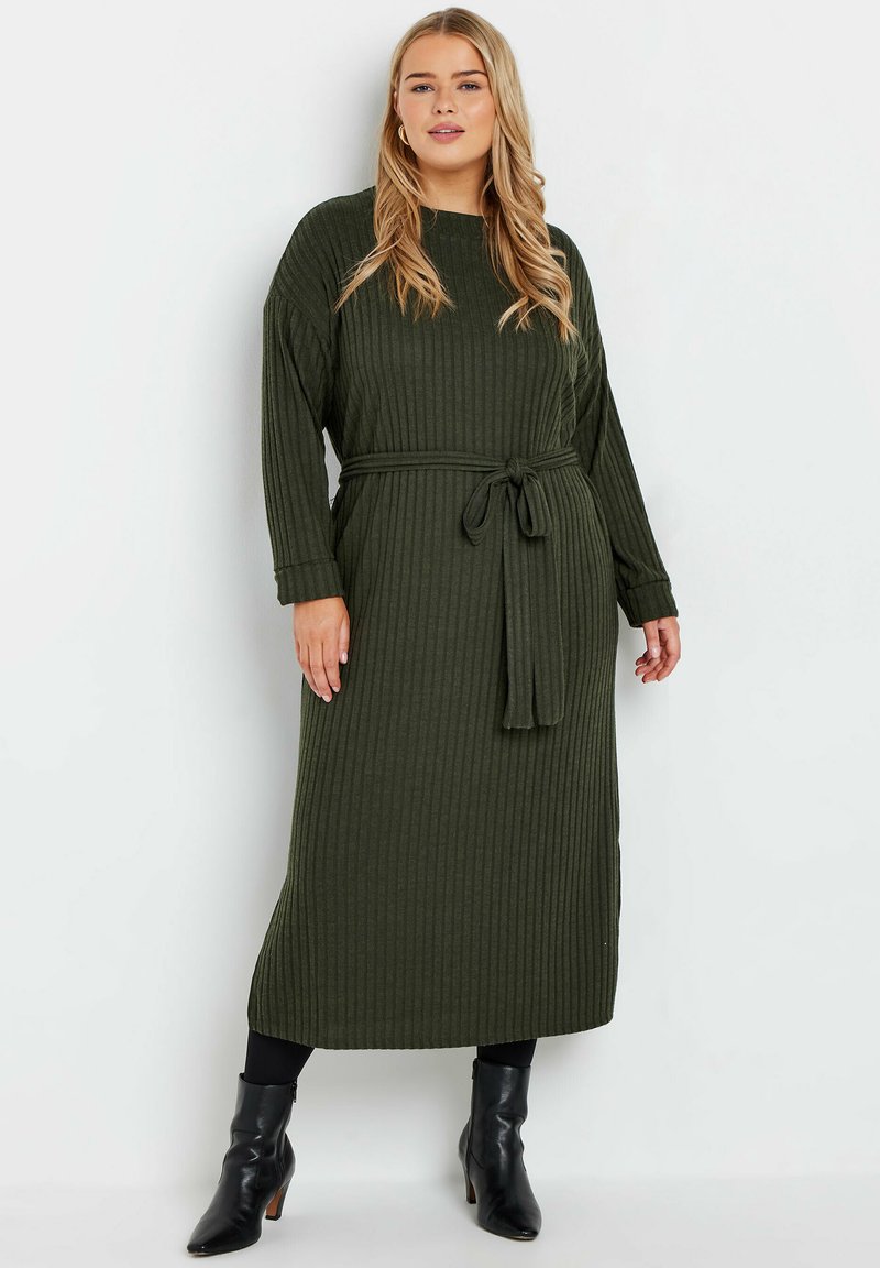 Robe longue verte à côtes avec des manches longues et une taille cintrée. Le tissu a une finition texturée. Portée avec des bottines noires.