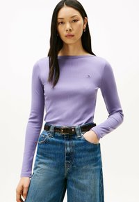 Helllila, geribbeltes Langarmoberteil mit einem Bootsausschnitt, kombiniert mit hochgeschnittenen blauen Jeans und einem schwarzen Gürtel mit einer silbernen Schnalle.