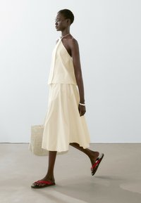 Krämfärgad ärmlös topp och kjolset, med en lös passform och lagerdesign. Accessoarerna inkluderar en vävd väska och röda flip-flops.