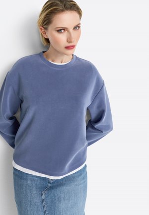 Sweatshirt bleu clair avec un col rond, coupe ample et poignets côtelés. Associé à un ourlet blanc superposé et une jupe en denim. Texture lisse.
