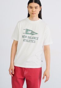 Camiseta blanca de algodón con un gráfico verde que dice "NEW BALANCE ATHLETICS" con un diseño de bandera, combinada con pantalones de mezclilla rojos.