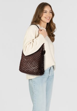 Femme souriante avec de longs cheveux portant un pull crème et un jean bleu clair, tenant un sac à bandoulière en cuir tissé marron foncé.