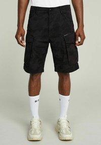 Czarne szorty cargo w kamuflażu z zamkami błyskawicznymi, wykonane z bawełny, z podwiniętym dołem i luźnym krojem, połączone z białymi skarpetkami i brązowymi trampkami.