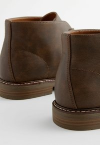 Botas de tobillo de cuero marrón con una textura suave, que presentan una punta redondeada, costuras en contraste y un tacón de madera apilada.