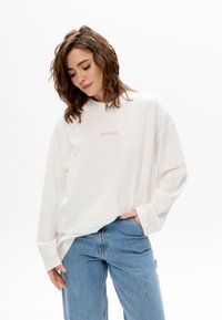 Weißes langärmliges Hemd mit Rundhalsausschnitt, verziert mit rosa gesticktem Text "MIRONS." Kombiniert mit hellblauen Denim-Jeans in lockerer Passform.