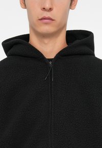 Haut en polaire noir avec un extérieur texturé, une capuche et une fermeture éclair à l'avant. Il dispose d'un cordon de serrage au niveau du cou pour un ajustement.