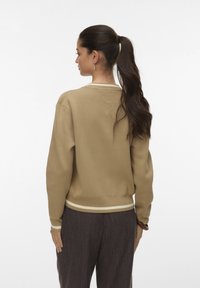 Vrouw met een donkere paardenstaart, gekleed in een beige sweater met witte afwerking en donkere gestreepte broek, staand met de rug naar de camera tegen een effen achtergrond.