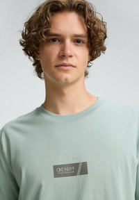 Jasnozielona koszulka z krótkim rękawem wykonana z bawełny, z teksturowanym nadrukiem graficznym zawierającym napisy "DENIM" oraz "07" w szarych i białych akcentach.