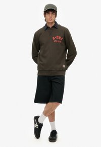 Oliwkowy sweatshirt z nadrukowanym logo, czarne szorty i czarne buty, w połączeniu z szarym czapką i białymi skarpetkami. Zawiera szczegół w postaci kratkowanego kołnierza.