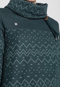 Dunkelgrüner Pullover mit hohem Kragen, der ein geometrisches Muster in hellgrün aufweist, mit Kordelzugdetails und einem kleinen Branding-Element.