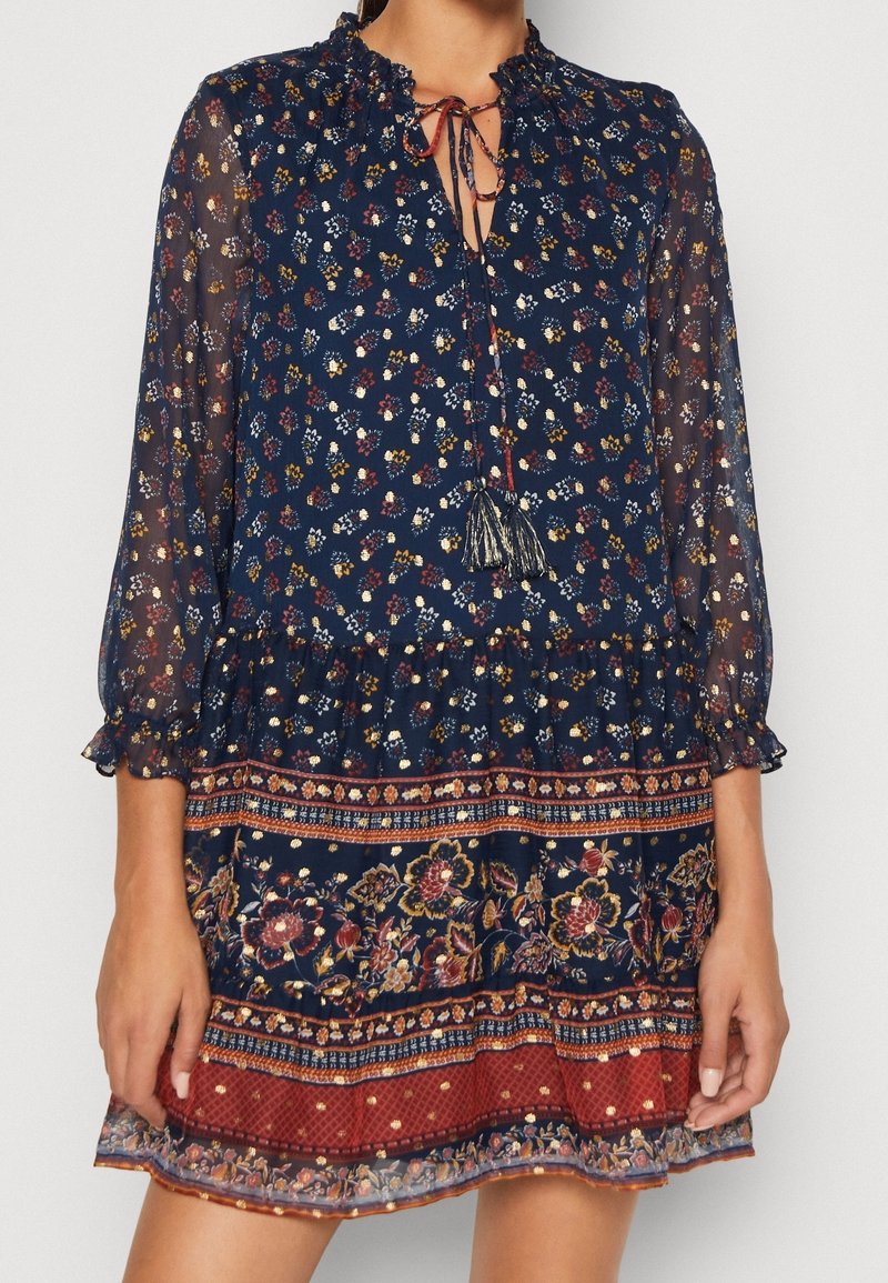 Marinblommig klänning med genomskinliga långa ärmar, volangkrage och kvastband. Har en mönstrad fåll i burgunder och guldaccenter.