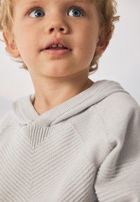 Sudadera con capucha de punto gris claro con un patrón texturizado, detalles acanalados y una capucha redondeada, colocada alrededor del cuello y los hombros de un niño.