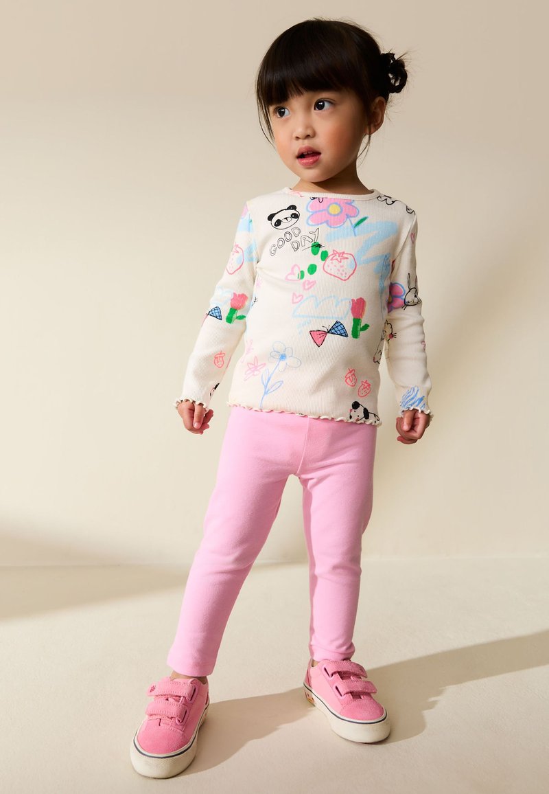 Junges Mädchen steht und trägt ein weißes Langarmshirt mit bunten Zeichnungen, pinke Leggings und pinke Turnschuhe vor einem einfarbigen Hintergrund.