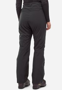 Person, der schwarze Skihosen und dunkelviolette Skischuhe trägt, von hinten vor einem einfarbigen weißen Hintergrund gezeigt.