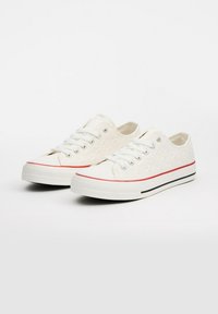 Zapatillas de lona blancas con textura decorativa, rayas rojas y negras en la suela, y ojales plateados para los cordones. Diseño de caña baja.