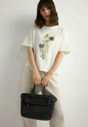 Młoda kobieta w białej koszulce z nadrukiem w groszki i jasnych spodniach, trzymająca czarną torbę na tle jednolitego tła.