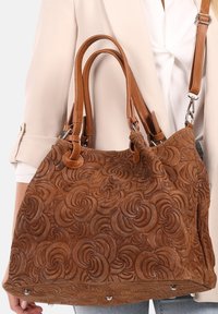 Bruine suede handtas met een gegraveerd, draaiend bloemenpatroon, dubbele leren handvatten en een afneembare schouderband. Accenten van zilverkleurig hardware.