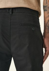 Pantalons noirs avec une texture lisse, avec une poche arrière à fermeture à bouton et une poche latérale discrète.