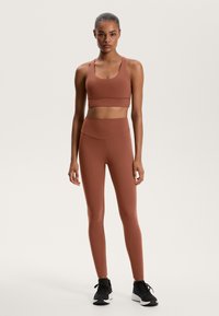 Rjav atletski crop top in visokorasle legice, izdelani iz gladkega, raztegljivega materiala, v kombinaciji s črnimi tekaškimi čevlji.