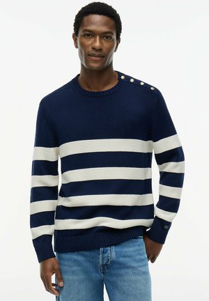 Mężczyzna ubrany w granatowy sweter w białe poziome paski z złotymi guzikami na ramieniu, w połączeniu z jasnoniebieskimi jeansami, stoi na białym tle.