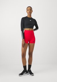 Crni dugi crop top s bijelim prugama, crvene kratke hlače visokog struka i crne sportske cipele s bijelim detaljima.