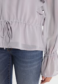 Blusa di un leggero lavanda con vita cin cin, dettagli arricciati e maniche a sbuffo elastiche. Realizzata in tessuto trasparente e leggero.