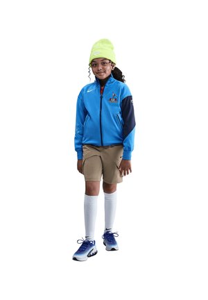Lächelndes Kind, das eine blaue Sportjacke, beige Shorts, weiße knielange Socken, blaue Turnschuhe und eine neongrüne Mütze trägt.