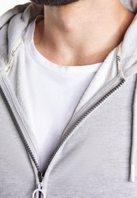 Sweat à capuche gris clair avec fermeture éclair, doublure intérieure blanche douce, zip argenté et un t-shirt blanc col rond en dessous.