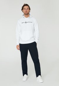 Sudadera blanca de algodón con bolsillo frontal y el logo "SAIL RACING", combinada con pantalones azul marino oscuros y zapatillas blancas, de pie sobre un fondo liso.