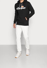 Sudadera negra con logo blanco y detalle en naranja, con un bolsillo frontal, combinada con pantalones blancos y zapatillas grises.