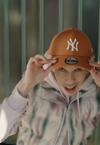 Bronzefarbene New York Yankees Cap im Fitted-Stil. Das Modell trägt eine leichte Steppjacke mit einem bunten, schimmernden Muster und einem weißen Hoodie darunter.