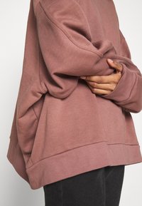 Sweat-shirt oversize à manches longues de couleur rose pâle ; confectionné en tissu doux avec un ourlet côtelé et de larges poignets. Poches latérales visibles.