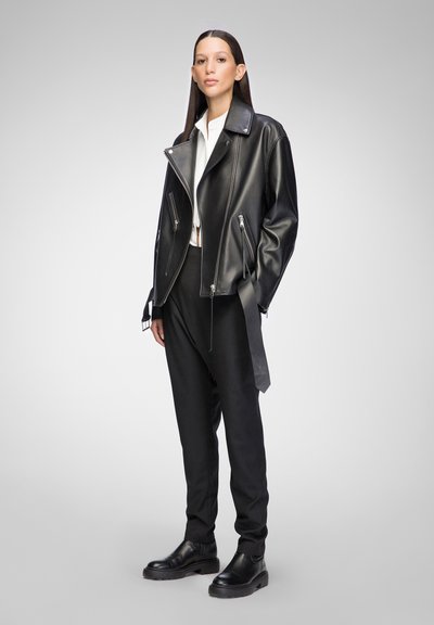 Schwarze Leder-Bikerjacke mit silbernen Reißverschlüssen, strukturierter Kragen und satinartigem Finish, kombiniert mit schwarzen, maßgeschneiderten Hosen und klobigen Stiefeln.