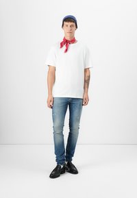 Camiseta blanca de manga corta, jeans azules claros desgastados, bandana roja alrededor del cuello, gorro azul de punto, zapatos negros tipo slip-on. Conjunto simple y casual.