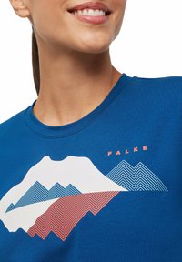 T-shirt in cotone blu con grafica di una montagna bianca decorata con schemi a zigzag blu e rossi. Il nome del marchio "FALKE" è stampato sopra il design.